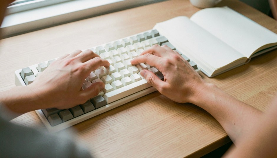 The two hands of one typist resting lightly on the keys of a white keyboard with ivory and grey keycaps, seen from behind the typist's shoulder, forearms over a light oak desk beside an open notebook with empty pages, soft daylight from a window